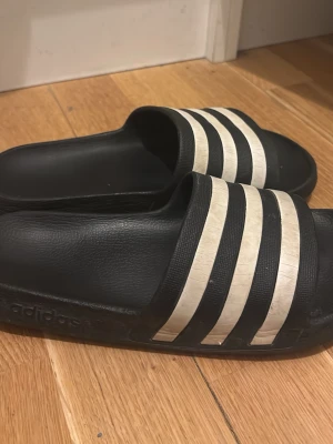 Svarta Adidas sandaler med vita ränder - Klassiska svarta sandaler från Adidas med tre vita ränder över foten. Tillverkade i syntetmaterial med platt sula och öppen tå. Perfekta för sommaren och enkla att bara kliva i.