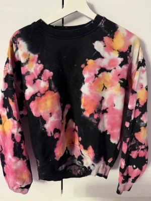  sweatshirt i rosa och svart - Unik tie dye sweatshirt med lång ärm i svart, rosa, gul och vit. Tröjan har rund halsringning och ribbade muddar vid ärmslut och nederkant. Tillverkad i mjuk bomull och polyesterblandning för skön känsla. Perfekt för dig som gillar färgstarka plagg och streetstyle.