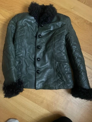 Mörkgrön skinnjacka  - Snygg mörkgrön skinnjacka från Nekhii med svart päls på krage, ärmslut och insida. Jackan har knappar framtill, två fickor och quiltade detaljer på ärmarna. Perfekt för dig som vill ha en unik och varm jacka med cool vintagekänsla.