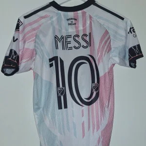 Inter Miami Messi fotbollströja Adidas - Säljer en Inter Miami fotbollströja med Messi 10 på ryggen. Tröjan är vit med ljusblå och rosa grafiska detaljer, svart krage och svarta detaljer. Adidas-logga och MLS-märke finns på bröstet. Materialet är lätt och andas, perfekt för match eller träning.