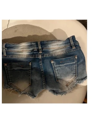 Blå jeansshorts med fransig kant - Snygga blå jeansshorts med slitna detaljer och fransig kant. Shortsen har klassiska bakfickor och en lågmidjad passform. Perfekta för varma dagar och en avslappnad stil.