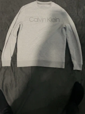 Grå sweatshirt från Calvin Klein - Snygg grå sweatshirt från Calvin Klein med logga tryckt framtill. Tröjan har rund halsringning, långa ärmar och ribbade muddar vid ärmslut och nederkant. Perfekt för en avslappnad och stilren look.