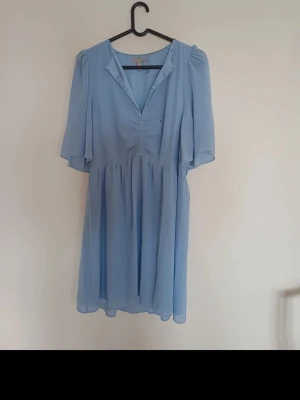 Ljusblå kortklänning från H&M - Supersöt ljusblå klänning från H&M med v-ringning och korta, fladdriga ärmar. Klänningen har en avslappnad passform och lätt rynkade detaljer framtill. Perfekt för varma dagar och enkel att styla med sneakers eller sandaler.