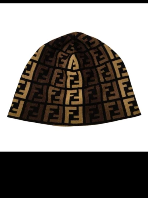Fendi beanie med FF-logga brun/guld - Snygg beanie från Fendi i brunt och guld med ikoniskt FF-monogram över hela mössan. Mjuk och stretchig passform, perfekt för att lyfta din streetstyle. Materialet är stickat och ger en lyxig känsla. Köptes för 355€.