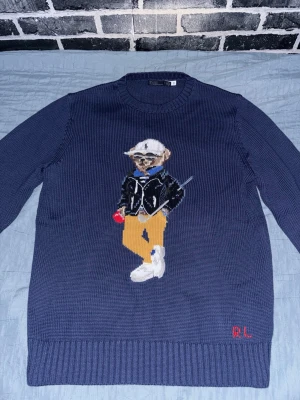 Ralph Lauren stickad tröja Polo Bear - Mörkblå stickad tröja från Ralph Lauren med ikoniska Polo Bear-motivet framtill. Björnen har vita sneakers, gula byxor, svart jacka och vit keps. RL-broderi i rött vid nederkanten. Rund halsringning och ribbade muddar. Perfekt för dig som gillar streetstyle och klassiska detaljer. Perfekt nu i vintern och inför våren. Storlek L men passar M väldigt bra. 