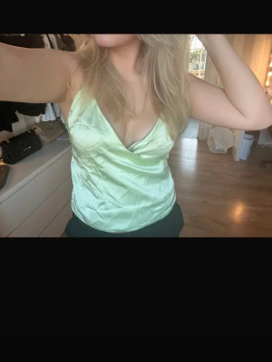 Ljusgrön omlott-linnetopp i satin - Snygg ljusgrön linne-topp med omlott och tunna axelband. Toppen har en glansig satin-finish och djup v-ringning framtill. Perfekt för varma dagar och ger en fräsch vibe till din outfit.