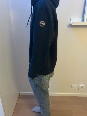 Mörkblå hoodie från Colmar - Snygg mörkblå hoodie från Colmar med klassisk huva och dragsko. Tröjan har en  Colmar-logga på ärmen och ribbade muddar vid ärmslut och nederkant. Perfekt för en avslappnad och stilren look. Killen på bilden är 176cm lång.