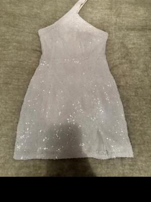 Vit glittrig oneshoulder klänning - Snygg vit kortklänning med glittriga paljetter och oneshoulder-design. Klänningen har en figurnära passform och en liten slits framtill. Perfekt för dig som vill sticka ut med en trendig och elegant look.