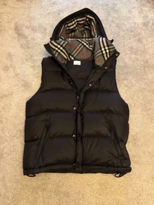 Svart dunjacka med avtagbara ärmar - Svart puffer dunjacka med avtagbara ärmar och huva. Insidan av huvan har ett klassiskt rutigt mönster i grått, vitt och rött. Jackan har dragkedja och knappar framtill samt två snedställda fickor med dragkedja. Snygg burberry jacka säljs för billigt bara dessa 3 dagar eftersom jag behöver pengar finns kvitto också till den passar både L och M knappt använd 