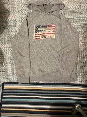 Grå hoodie från Ralph Lauren - Snygg grå hoodie från Ralph Lauren med ett stort patch framtill som har amerikansk flagga och texten 'Denim & Supply'. Hoodien har långa ärmar och klassisk huva. Perfekt för en avslappnad och cool stil.