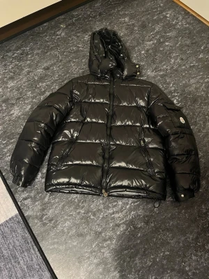 Svart pufferjacka med huva Moncler - Säljer en svart, glansig pufferjacka från Moncler med huva och dragkedja framtill. Jackan har quiltad design, lång ärm och logotyp på ärmen. Perfekt för kalla dagar och riktigt snygg streetstil.