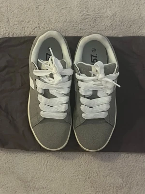 Gråa sneakers med vita detaljer - Stilrena grå sneakers med vita snören och vit sula. Klassisk låg modell med rund tå och platt sula. Perfekta för en avslappnad och trendig look. Köpt från Lager 157, använt en gång 🩶