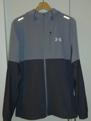 Grå vindjacka från Under Armour - Snygg tvåfärgad vindjacka från Under Armour i ljus- och mörkgrått. Jackan har huva, hel dragkedja framtill, reflekterande detaljer på axlarna och logga på bröstet. Perfekt för blåsiga dagar och sportiga outfits.
