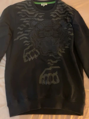 Svart sweatshirt med tiger från Kenzo - Cool svart sweatshirt från Kenzo med ikoniskt tigertryck och mönster i svart på bröstet. Tröjan har rund halsringning och ribbade muddar. Perfekt för dig som gillar streetwear och vill sticka ut med en unik design.