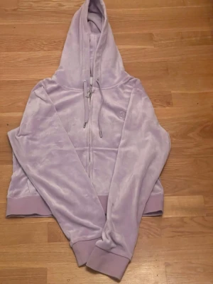 Syrenlila velour hoodie med dragkedja från Juicy Couture  - Säljer en ljuslila hoodie från Juicy Couture i mjuk velour. Älskar hoodien men jag  har inte lyckats hittat byxorna i samma färg och modell så får sälja den. Hoodien har dragkedja framtill, fickor och justerbar huva med snören. Ribbstickade muddar vid ärmslut och nederkant ger en snygg passform. Perfekt för en chill och trendig look.