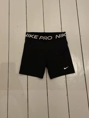 Svarta Nike Pro träningstights shorts - Svarta tighta träningstights shorts från Nike Pro med bred resår i midjan där det står 'NIKE PRO' i vitt. Klassisk vit Nike-logga på ena benet. Perfekta för gym, löpning eller annan träning. Stretchigt material för maximal rörelsefrihet.