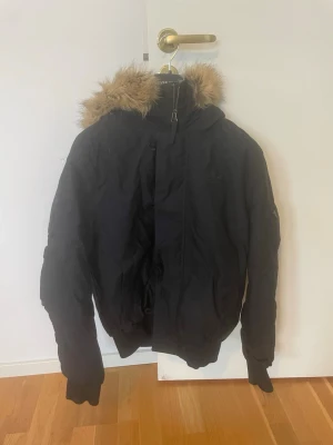 Svart parkas med fuskpäls - Svart parkas med huva och brun fuskpäls runt kragen. Jackan har dragkedja framtill och långa ärmar med muddar. Perfekt för kalla dagar och har en klassisk, enkel design.