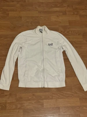 Vit ziptröja från EA7 Emporio Armani - Snygg vit ziptröja från EA7 Emporio Armani med hög krage och dragkedja framtill. Tröjan har två fickor på sidorna och en diskret logga på bröstet. På ryggen finns svarta ränder upptill. Perfekt för en clean och sportig look.