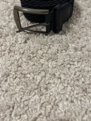 Svart flätat bälte med metallspänne - Snyggt svart flätat bälte med silverfärgat metallspänne och detaljer i läderimitation. Bältet har en stilren design och passar perfekt till jeans eller chinos. Enkel att justera tack vare det flexibla materialet.