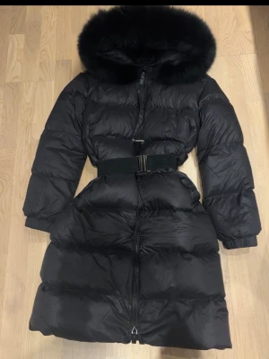 Svart lång dunjacka med bälte och luva - Snygg svart lång dunjacka med stor luva och fluffig pälskant. Jackan har dragkedja framtill och ett brett bälte i midjan för extra form. 