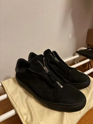 Svarta arigatos med dragkedja i mocka - Svarta arigato sneakers i mocka med cool dragkedja framtill istället för klassisk snörning. Snygg minimalistisk design med rund tå och platt sula. Perfekta för dig som gillar stilrena och moderna skor.Det enda är att sulan är lite sliten se bild 5 men det går att fixa. Annars är skon fel fri. Du får med dig arigato lådan och även dust bagen.