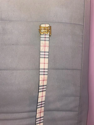 Rutigt bälte med guldspänne Burberry - Snyggt bälte från Burberry med klassiskt rutmönster i beige, rosa och svart. Bältet har ett guldfärgat metallspänne med Burberrys logga. Perfekt accessoar för att lyfta din outfit och ge en exklusiv touch. Tillverkat i slitstarkt material.
