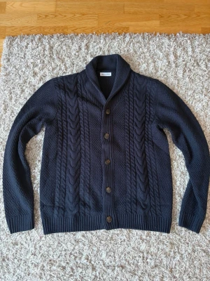 Stickad kofta med knappar från Jack N Jones - Två stilrena stickade koftor från Jack N Jones, en i mörkblått och en i beige. Båda har grovt kabelstickat mönster, ribbade muddar och knäppning framtill med fem knappar. Perfekt för lager-på-lager och en klassisk look. 299 kr styck och 500 kr som paketpris för båda!