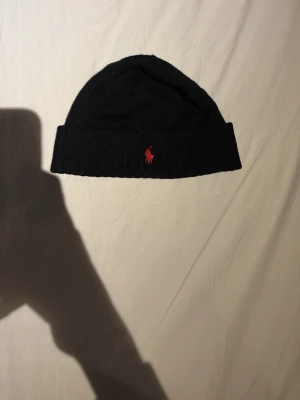 Svart mössa från Polo Ralph Lauren - Klassisk svart ribbstickad mössa från Polo Ralph Lauren med uppvikt kant och röd broderad logga framtill. Perfekt accessoar för kyliga dagar och enkel att matcha med olika stilar.