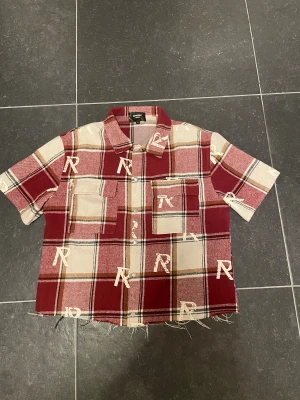 Rutig kortärmad skjorta Represent - Kortärmad skjorta från Represent med röd, beige och brun rutig design. Skjortan har tryckta R-loggor, två bröstfickor och rå, fransig nederkant. Tillverkad i mjuk bomull och har knappar framtill. Perfekt för dig som gillar streetwear vibes. Klippt längst ner för att ha en mer boxig look