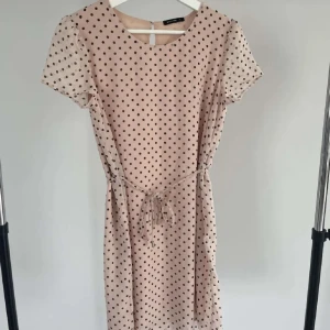 Beige prickig kortklänning  - Säljer en beige kortklänning från Selected Femme med svarta prickar, rund hals och korta ärmar. Klänningen har ett knytband i midjan för extra snygg passform och är tillverkad i ett lätt och luftigt material. Perfekt för dig som gillar en söt och trendig look.