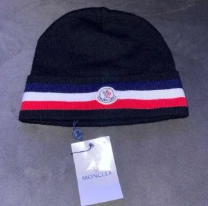Svart Moncler mössa med ränder - Svart mössa från Moncler med klassiska blå, vita och röda ränder runt kanten. Mössan har Moncler-logga framtill och är stickad i mjukt material. Perfekt accessoar för att hålla stilen under kyliga dagar.