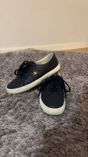 Marinblå Ralph Lauren skor med snörning - Snygga marinblå Ralph Lauren sneakers i skinn med en vit platt sula med snörning. Perfekt till dig som gillar en enkel men stilren sneaker. Nypris 1245kr
