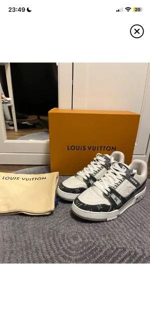 Louis Vuitton grå/vita sneakers - Louis Vuitton sneakers i vit och grå färg med klassiskt monogrammönster. Låg modell med snörning och platt sula. Skorna har detaljer i skinn och textil, samt tydliga LV-loggor på sidan och sulan. Snygg kontrast mellan ljusa och mörka toner, perfekt för dig som gillar exklusiv streetwear.
