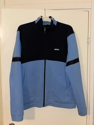 Blå och marinblå ziptröja från BOSS - Snygg ziptröja från BOSS i ljusblå och marinblå färg. Tröjan har hel dragkedja, hög krage och långärmade ärmar. På armarna finns ett marinblått band med BOSS-logga. Perfekt för en chill och sportig look.