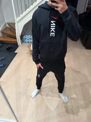 Nike tracksuit - Svart hoodie från Nike i storlek XS med stor vit logga och röd swoosh på bröstet. Tröjan har huva med snörning, magficka och långa ärmar. Materialet är mjukt och känns som bomull och polyester, perfekt för chill eller träning. Går att köpa separat, varsin köpt för 800kr hos Jd Sports, 1600kr totalt