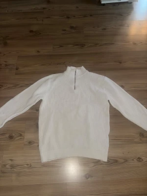 Vit stickad tröja med half zip från Smog - Säljer en vit stickad tröja från Smog i regular fit. Tröjan har en ribbad struktur, hög krage och snygg half zip-dragkedja framtill. Perfekt för dig som gillar enkel och clean stil med lite edge. Långärmad och passar till många olika looks.