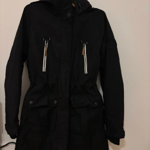 Svart parkajacka från McKinley - Svart parkajacka från McKinley med huva, två stora fickor med lock och två bröstfickor med vita dragkedjor och läderdetaljer. Jackan har justerbar midja och är något stor i storleken. 