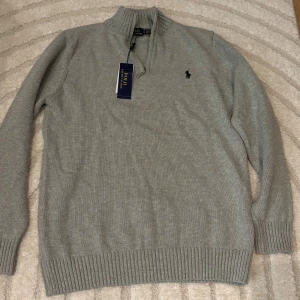 Grå stickad half zip från Polo Ralph Lauren - Snygg grå stickad tröja från Polo Ralph Lauren med half zip och ribbade muddar. Klassisk broderad logga på bröstet och hög krage. Perfekt för dig som gillar stilrena och tidlösa plagg med premiumkänsla. Inte äkta med prislapp osv med!