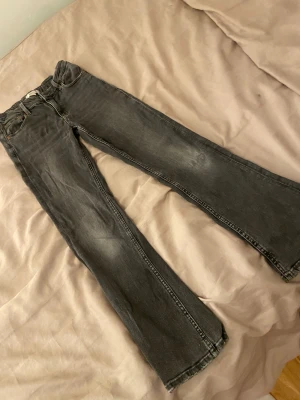 Grå bootcut jeans från Gina Tricot - Snygga grå jeans från Gina Tricot i bootcut-modell med klassisk femficksdesign och slitna detaljer vid benslut. Jeansen har knappstängning och bälteshällor, tillverkade i mjukt jeanstyg som ger en cool vintagekänsla. Jeansen har ett igensytt hål på ena knät men syns knappt.💕