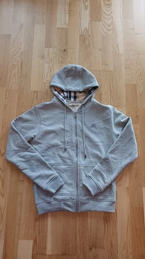 Grå hoodie från Burberry med dragkedja - Säljer en grå hoodie från Burberry med klassiskt rutigt foder i huvan och längs insidan. Tröjan har dragkedja framtill, snörning i huvan och ribbade muddar. Perfekt för dig som gillar stilrena och exklusiva plagg med en twist.