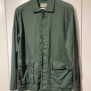 Grön overshirt från East West - Snygg grön overshirt från East West i storlek S. Jackan har klassisk krage, stora fickor framtill och knäppning med knappar hela vägen. Perfekt för lager-på-lager och chill stil. Materialet är bomull och modellen är långärmad.