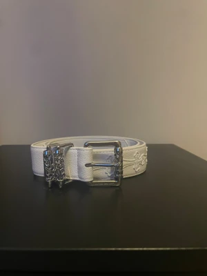 Vitt skärp med silverdetaljer - Snyggt vitt bälte i skinn med silvrigt spänne och dekorativa detaljer. Bältet har broderade kors längs remmen och ett unikt, mönstrat metallspänne som ger en cool touch till outfiten. Perfekt accessoar för att lyfta din stil.