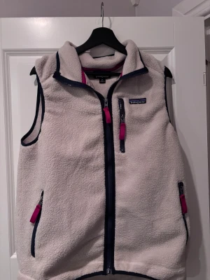 Patagonia väst  - Säljer en beige fleeceväst från Patagonia i storlek M. Västen har marinblå detaljer, dragkedjor med röda band och hög krage. Utrustad med tre fickor med dragkedja och ett broderat Patagonia-märke på bröstet. Perfekt för lager-på-lager och chill utomhus.Helt ny aldrig använd.