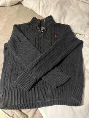 Svart stickad tröja Polo Ralph Lauren - Svart kabelstickad tröja från Polo Ralph Lauren med röd broderad logga på bröstet. Tröjan är textilfärgad också då den har varit solblekt men ser fortfarande riktigt bra ut, vid fundering och frågor är det bara säga till och priset går självklart att diskuteras!😊🤗