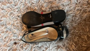 Burberry ballerinaskor med klassiskt mönster - Snygga ballerinaskor från Burberry i beige och svart med klassiskt rutigt mönster och röda detaljer. Skorna har svarta lackdetaljer, dekorativt spänne framtill och platt sula. Perfekta för dig som gillar en tidlös och lyxig stil. Osäker på äkta men ser ut att vara det