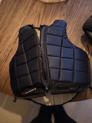 Svart quiltad säkerhetsväst Racesafe - Säljer en svart quiltad säkerhetsväst från Racesafe, perfekt för ridning. Västen har rutmönster, dragkedja framtill och är tillverkad i polyester med nylon och elastan i fodret. Snygg och funktionell design som ger bra skydd vid aktivitet.