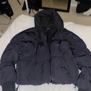 Svart pufferjacka från H&M med huva - Säljer en svart pufferjacka från H&M med huva och dragkedja. Jackan är vadderad och har breda muddar vid ärmsluten. Tillverkad i 100% polyester, perfekt för kalla dagar. Enkel och stilren design som funkar till det mesta.