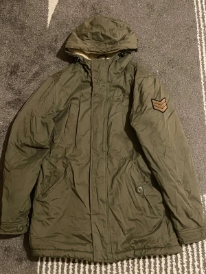 Olivgrön parkajacka med fuskpälsfoder - Olivgrön parkajacka från SMOG med mysigt fuskpälsfoder och quiltad insida. Jackan har huva, flera fickor och en cool patch på ärmen. Perfekt för kalla dagar när du vill hålla stilen och värmen.