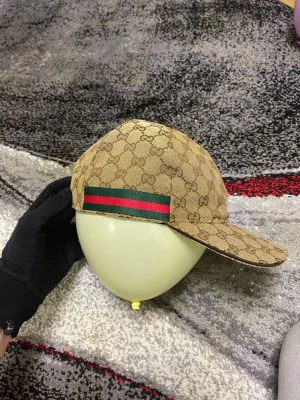 Gucci beige keps med GG-mönster - Snygg beige keps från Gucci med klassiskt GG-mönster i brunt och en grön-röd rand på sidan. Justerbar rem baktill i mörkbrunt skinn. Perfekt för dig som vill ha en lyxig och trendig look. Materialet är slitstark bomull med detaljer i skinn.