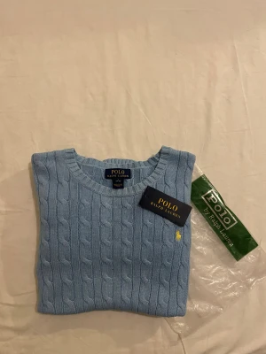 Ljusblå stickad tröja Polo Ralph Lauren - Snygg ljusblå stickad tröja från Polo Ralph Lauren med klassiskt kabelstickat mönster och gul broderad logga på bröstet. Rund hals och ribbade muddar vid ärmslut och nederkant. Perfekt för dig som gillar stilrena och tidlösa plagg.